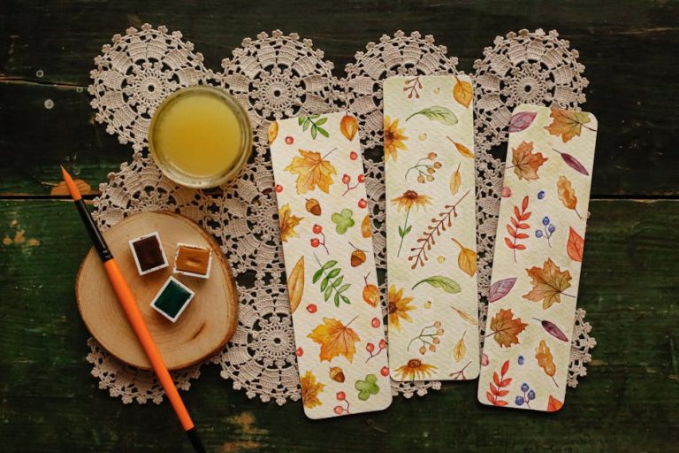 Artistic flat lay featuring watercolor paints, brush, and autumn-themed bookmarks on crochet doily.