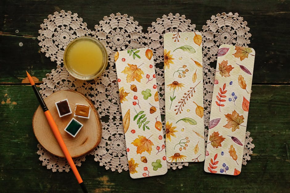 Artistic flat lay featuring watercolor paints, brush, and autumn-themed bookmarks on crochet doily.