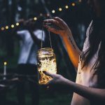 Crop anonymous female with long dark hair in casual clothes standing with glass jar with glowing garland in nature in evening time