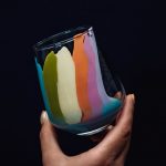 A close-up of a hand holding a glass with vibrant rainbow colors on a dark background.