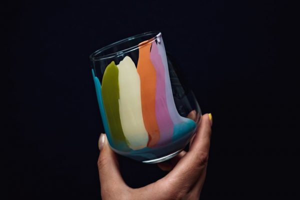 A close-up of a hand holding a glass with vibrant rainbow colors on a dark background.